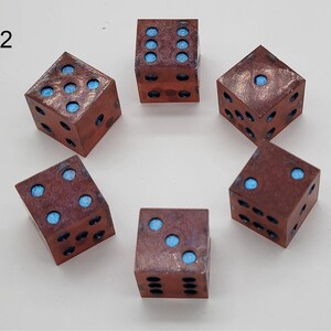 May include: Six square dice arranged in a circle, each featuring a metallic copper-toned finish with a weathered texture. The recessed pips on each face are filled with a bright, matte light blue pigment. The number two is printed in black.