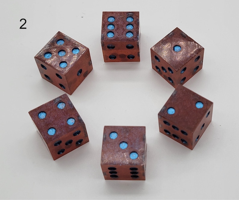 May include: Six square dice arranged in a circle, each featuring a metallic copper-toned finish with a weathered texture. The recessed pips on each face are filled with a bright, matte light blue pigment. The number two is printed in black.