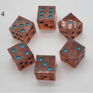 May include: Six metallic dice arranged in a circle, each featuring a weathered copper-toned finish with recessed circular pips filled with bright turquoise blue pigment. The number 4 is printed in black ink to the left of the dice arrangement.