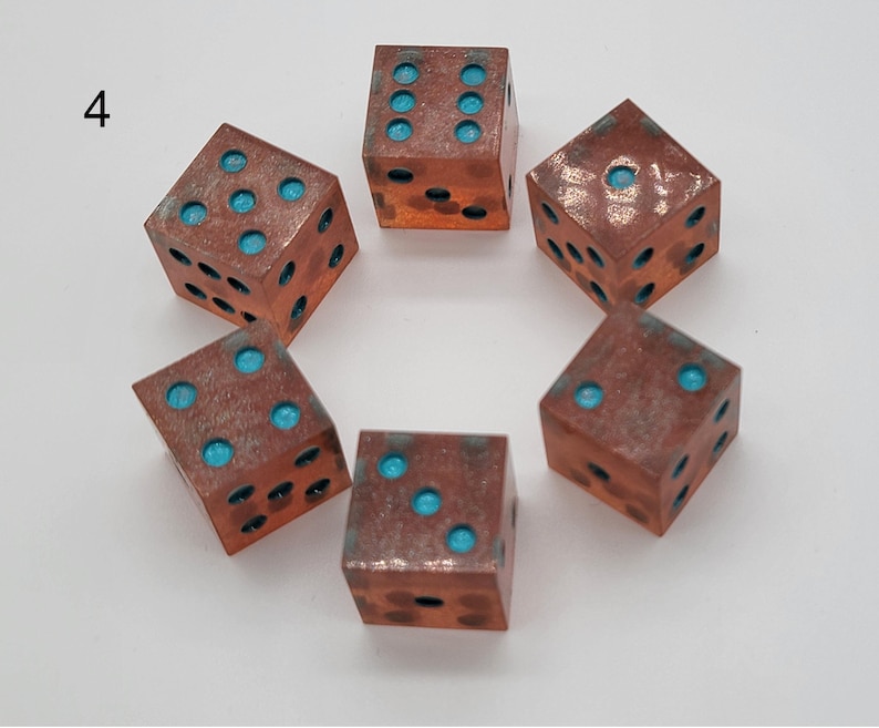 May include: Six metallic dice arranged in a circle, each featuring a weathered copper-toned finish with recessed circular pips filled with bright turquoise blue pigment. The number 4 is printed in black ink to the left of the dice arrangement.