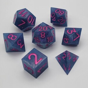May include: Set of seven polyhedral dice in shades of blue with pink numbering. The dice are in various geometric shapes, including a d4, d6, d8, d10, d12, d20, and a percentile die. Ideal for tabletop games.