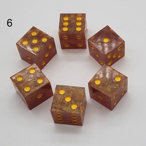 May include: Six square dice arranged in a circle, each featuring a translucent reddish-brown base with embedded gold-colored glitter and bright yellow pips. The number 6 is printed in black in the upper left corner of the image frame.