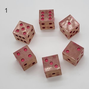 May include: Six six-sided dice arranged in a circle, each featuring a metallic copper-toned finish with recessed circular pips filled with a vibrant pink color. The number one is visible on the top face of the die located at the top right.