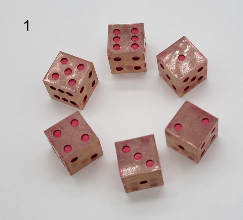 May include: Six six-sided dice arranged in a circle, each featuring a metallic copper-toned finish with recessed circular pips filled with a vibrant pink color. The number one is visible on the top face of the die located at the top right.