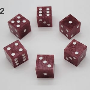 May include: Six six-sided dice arranged in a circle, each featuring a deep maroon, marbled resin finish with white pips. The number 12 is printed in black in the upper left corner. Each die displays different face values including one, two, three, four, five, and six.