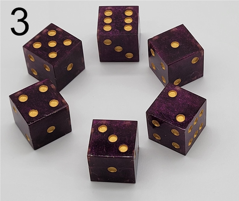 May include: A set of five six-sided dice in a deep purple color with a subtle glittery finish. Each die features recessed circular pips filled with a metallic gold paint. The number 3 is printed in black in the upper left corner of the image.