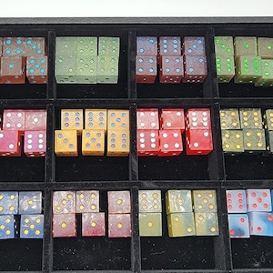May include: A collection of numerous translucent and opaque six-sided dice in various colors, including red, blue, green, yellow, and brown, organized into a black display tray with fifteen individual square compartments for storage and sorting.