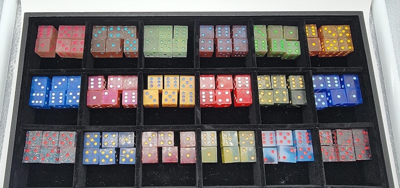May include: A collection of numerous translucent and opaque six-sided dice in various colors, including red, blue, green, yellow, and brown, organized into a black display tray with fifteen individual square compartments for storage and sorting.