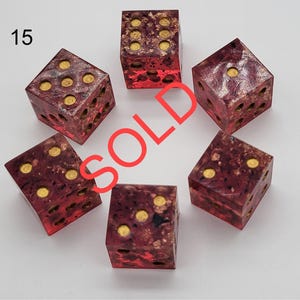 May include: A set of five six-sided dice with a translucent deep red and purple marbled resin body containing gold-colored flakes. Each die features circular gold-colored pips. The number 15 is printed in the top left corner and the word SOLD is overlaid.