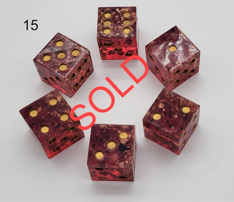 May include: A set of five six-sided dice with a translucent deep red and purple marbled resin body containing gold-colored flakes. Each die features circular gold-colored pips. The number 15 is printed in the top left corner and the word SOLD is overlaid.