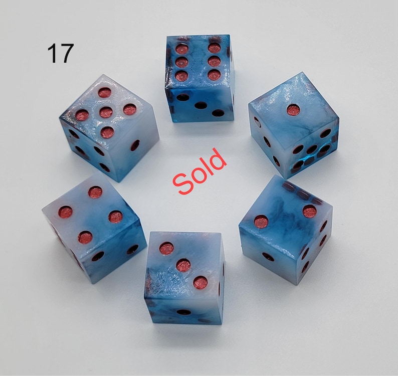 May include: Six translucent dice with a swirling blue and white marbled pattern and recessed red pips. The number 17 is printed in black in the upper left corner, and the word Sold is printed in red in the center of the arrangement.