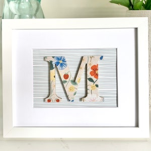 May include: A white frame with a white mat and a floral patterned letter M in the center. The letter is raised and has a gray and white striped background.