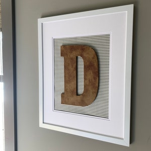 May include: A white picture frame with a brown letter "D" on a striped background.