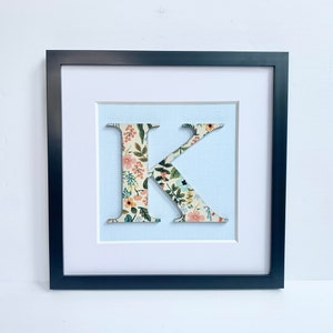 May include: A black framed picture with a white mat and a floral patterned letter K in the center. The letter is raised and appears to be made of fabric.