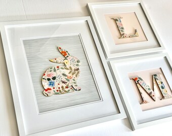 May include: Three white picture frames with a floral pattern. The frames are arranged in a row. The left frame features a floral patterned rabbit, the middle frame features the letter L, and the right frame features the letter M.