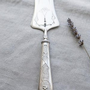May include: A silver-toned metal cake server featuring an elongated, triangular blade with engraved line detailing. The handle is cylindrical with a textured, geometric pattern and a small, circular floral motif embossed at the base of the handle.