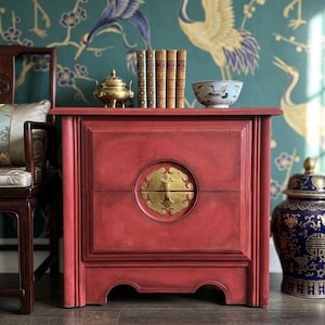 May include: A rectangular red wooden cabinet featuring a central circular brass plate with a vertical handle. The cabinet has a flat top surface holding a small brass incense burner, four vintage books with gold spines, and a white ceramic bowl.