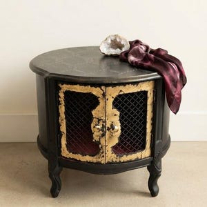 Peut inclure: Une table d'appoint ronde noire avec des pieds courbés et deux portes de placard dotées de cadres texturés à la feuille d'or, d'une grille métallique sombre et de boutons dorés. Une géode et un tissu bordeaux drapent le dessus.