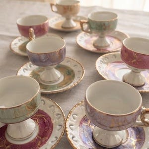 Peut inclure: Une collection de sept tasses à thé sur pied avec soucoupes assorties dans diverses couleurs pastel, notamment rose, violet et vert. Chaque tasse présente un intérieur blanc, un bord doré et une base sur pied, avec des soucoupes à motifs dorés.