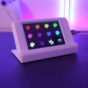 May include: A white, rectangular device with a small screen displaying colorful icons. The screen shows various palette and tool icons. The device is connected to a cable and sits on a dark surface, with a game controller in the background.