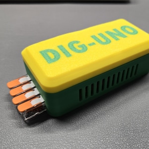 May include: A rectangular electronic device with a yellow top and green base. The text "DIG-UNO" is printed in green on the top. Several orange and white connectors protrude from one side. The device is on a gray surface.