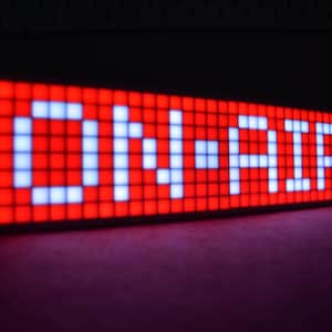 May include: A rectangular LED sign displaying the words "ON-AIR" in white against a red background. The sign is composed of individual illuminated squares, creating a pixelated effect. The sign is angled, with a dark background.