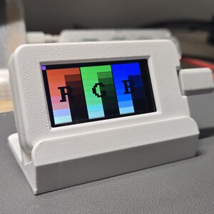 May include: A white plastic electronic display device featuring a rectangular screen that shows a color test pattern with the letters R, G, and B in black. The device is connected to a red power cable and sits in a matching white plastic stand.
