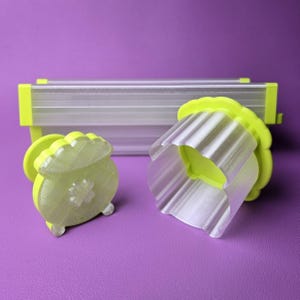 May include: Two lime green and clear plastic cookie cutters in the shape of a pot of gold and a scalloped edge. A clear plastic rectangular object with lime green accents is in the background. The objects are on a purple surface.