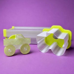 May include: A set of 3D-printed cookie cutters in the shape of a truck and a flower, with a clear plastic storage container. The truck is light yellow with transparent wheels. The flower is white and yellow. The background is purple.