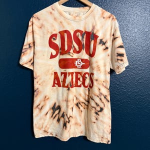 Peut inclure: T-shirt tie-dye crème et marron avec l'inscription rouge "SDSU AZTECS". Le t-shirt est suspendu à un cintre en bois sur fond bleu foncé. Le t-shirt a un motif.