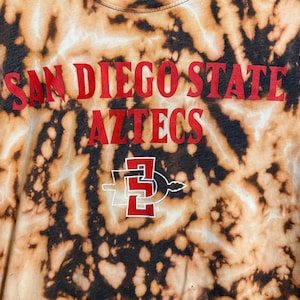 Peut inclure: T-shirt tie-dye avec les mots "SAN DIEGO STATE AZTECS" en lettres rouges. Le t-shirt présente également le logo des Aztecs de San Diego State en rouge et blanc. Le t-shirt a un motif tie-dye marron et noir.