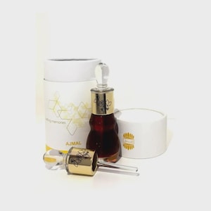 May include: A perfume bottle with a gold cap and clear glass applicator, next to a matching open bottle and two white cylindrical boxes. The bottle contains a dark amber liquid. The boxes have gold geometric designs and the word "AJMAL".