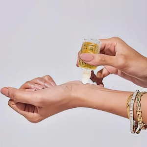 May include: A small, clear glass bottle with gold-colored liquid is being applied to a wrist. The bottle has gold decorative accents. The wrist has gold and silver bracelets. The background is a plain white.