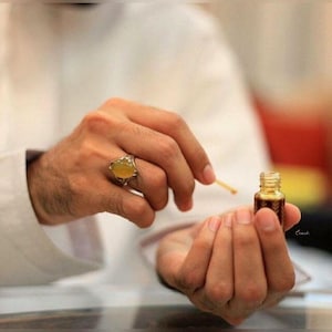 May include: A small, ornate glass bottle filled with a dark amber liquid is held between two hands. A gold ring with a large, yellow gemstone adorns one hand. A small wooden stick is held near the bottle, suggesting its use. The image evokes a sense of luxury and fragrance.