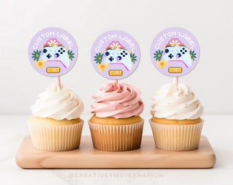 May include: Three cupcakes decorated with purple and white round labels featuring a cartoon video game controller with the text "Custom Label" and "Start".