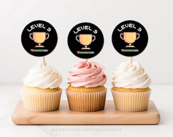 May include: Three cupcakes with black and white round toppers that say "Level Up" with a pixelated trophy and a progress bar. The cupcakes are on a wooden surface.