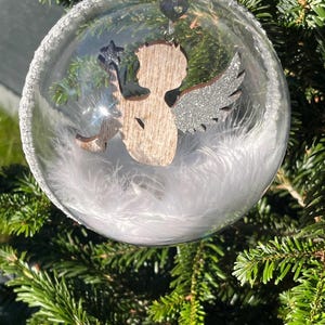 Puede incluir: Una bola de Navidad de cristal transparente llena de plumas blancas y una figura de ángel de madera. El ángel tiene alas de purpurina plateada y sostiene una estrella. La bola cuelga de un hilo plateado, rodeada de agujas de pino verdes.