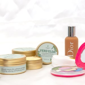Puede incluir: Una colección de productos de belleza que incluye un estuche compacto rosa con espejo, un pequeño tarro de bálsamo limpiador etiquetado como "VERD'ELDA CLEANSING BALM", una botella de base de maquillaje etiquetada como "DIOR" y un lápiz labial morado.