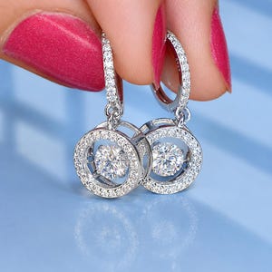 May include: A pair of silver-toned hoop earrings featuring a circular frame encrusted with small clear stones. Each earring has a central, suspended round clear stone that hangs within the circle. The metal is stamped with S925.