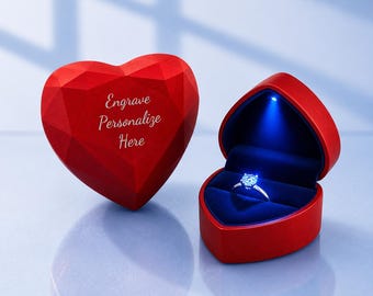 May include: A red heart-shaped ring box with a faceted exterior and the text Engrave Personalize Here, paired with an open matching box containing a silver ring illuminated by a small blue LED light inside the dark blue velvet interior lining.