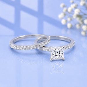 May include: A silver-toned engagement ring set featuring a princess-cut center stone and a matching band. Both rings are adorned with small, clear pavé-set stones along the shank, creating a cohesive and sparkling appearance for this jewelry set.