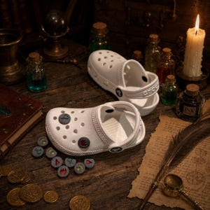 May include: Two white Crocs clogs with decorative charms, displayed on a wooden surface with other fantasy-themed items. The charms are round with various symbols. A lit candle, potion bottles, and coins are also visible.