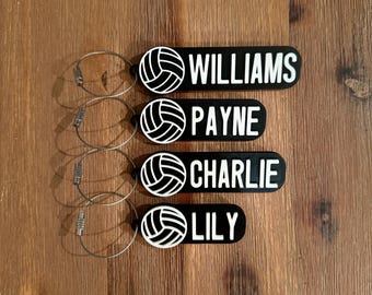 May include: Four black and white volleyball keychain tags with silver key rings. The tags have a volleyball graphic and the names "WILLIAMS", "PAYNE", "CHARLIE", and "LILY" printed in white.