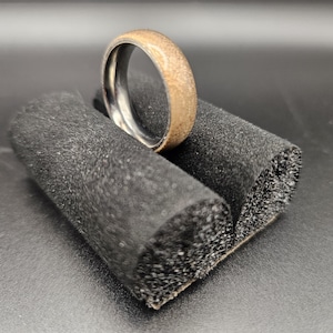 May include: A brown wooden ring with a silver band, displayed on a black foam ring display stand.