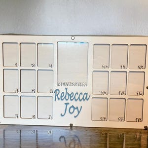 May include: A rectangular off-white wooden photo frame featuring eighteen small rectangular slots numbered 1 through 18, a larger central vertical slot, and the laser-cut text NEWBORN Rebecca Joy in blue script lettering on the front surface.