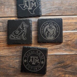 Puede incluir: Cuatro posavasos cuadrados de pizarra negra con diseños grabados. Uno presenta el contorno de Texas con "A&M", otro un perro con "A&M", un tercero con un hombre y "Texas A&M 12th Man", y el último con "Texas A&M EST:1876".