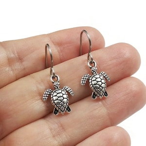 May include: Silver dangle earrings featuring a small turtle charm. The earrings are on a hand.