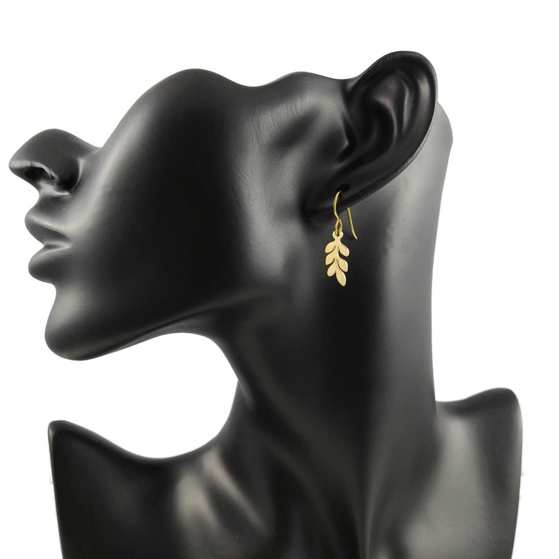 May include: Gold leaf dangle earrings with a delicate design. The earrings are shown on a black mannequin head.