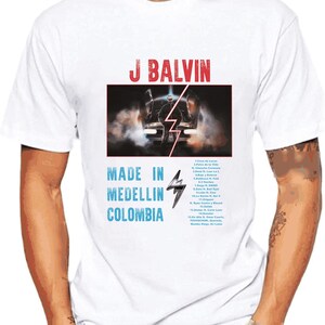 Pode incluir: Camiseta branca com uma estampa gr&aacute;fica de J Balvin, um carro e um raio. O texto "Made in Medellin, Colombia" est&aacute; impresso abaixo do gr&aacute;fico. A lista de faixas do &aacute;lbum "Jose" est&aacute; impressa no lado direito da camiseta.