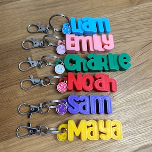May include: Keychains with names Liam, Emily, Charlie, Noah, Sam, and Maya in various colors. Each keychain has a smiley face charm and a silver clasp.